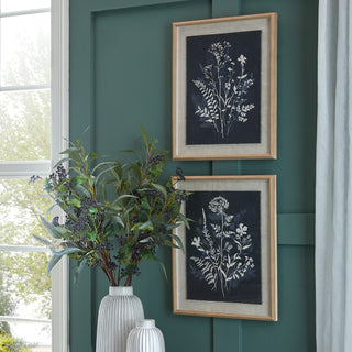 Two framed botanical prints hung on a dark green panelled wall above a white vase of dark foliage.