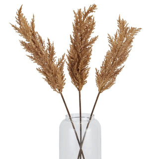 Grouping of three brown faux pampas grass stems arranged in a simple glass vase.