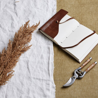 Faux brown pampas grass stem styled on white linen, beside a leather notebook and metal shears on a brown surface.