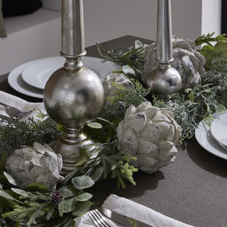 Ceramic artichoke ornaments decorating a dining table centrepiece with silver candle holders and winter greenery.