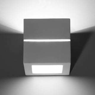 Front view of the cubic ceramic wall lamp showing symmetrical light distribution upwards, downwards, and through the central slot.