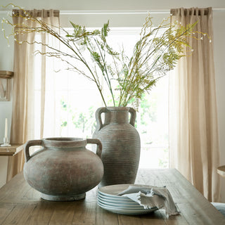 Rustic urn displayed next to a taller vase filled with foliage, on a wooden dining table.