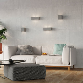 Grey ceramic wall light installed in a styled lounge setting above a white sofa on a light grey tiled wall.