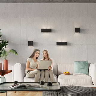 Four black wall lamps, cubic and rectangular variants, illuminating a grey tiled wall above a sofa.