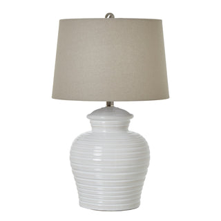Front-on view of a white ceramic table lamp with a ribbed base and a tapered beige linen drum shade on a white background.