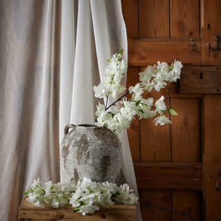 White artificial cherry blossom stems in a rustic grey pot on a wooden crate against sheer fabric and wooden panelling.