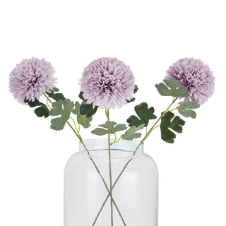 Three light purple chrysanthemum stems with green leaves in a clear glass vase.