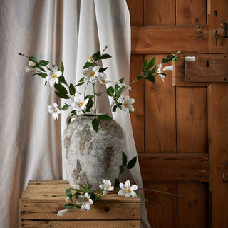 Artificial clematis stems in a large rustic vase on a wooden crate, set against white fabric and a wooden door.