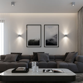 Two concrete ceiling fixtures illuminating a large grey sofa and framed artwork, alongside matching wall lights.