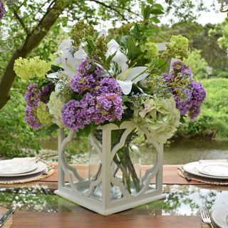 Artificial cream gardenia mixed with purple hydrangeas and white lilies in a vase on an outdoor dining table beside a stream.