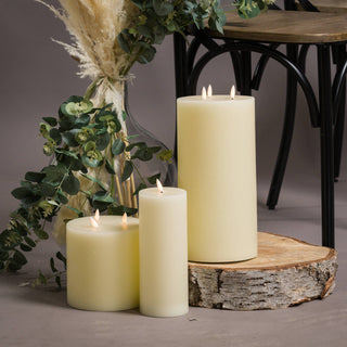 Three cream LED pillar candles of different sizes, one on a log slice, styled with eucalyptus and pampas grass.