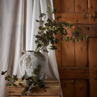 Variegated maple stem arranged in a large textured vase, set against sheer curtains and rustic wooden panelling.