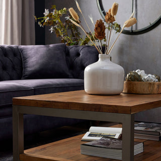 The dried bouquet displayed in a white vase on a wooden table, with a dark tufted sofa behind.
