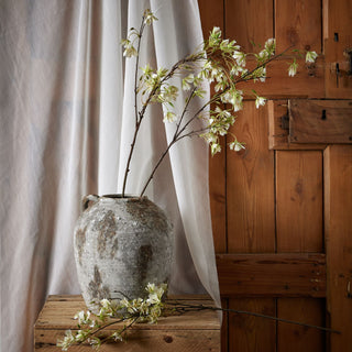 White flowering stems in a textured grey vase on a wooden crate, set against a sheer drape and wood panelling.