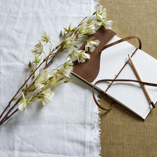 Styled flat lay showing the white-flowered stem on white linen beside an open sketchbook.