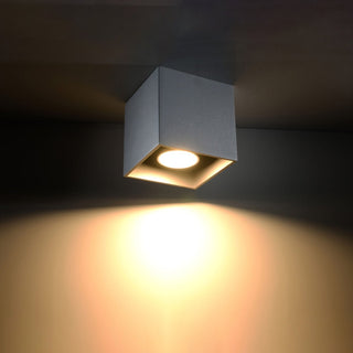 Grey cubic ceiling light fixture switched on, casting a warm, bright spotlight beam onto the surface below.