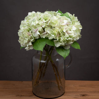 A bunch of artificial green and white hydrangeas in a large, dark glass vase on a wooden surface.