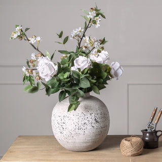 Eucalyptus filler mixed with artificial white roses and flowering branches in a large round vase on a rustic wooden table.