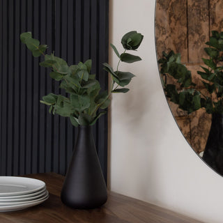 Artificial eucalyptus in a black vase on a wooden surface, next to a round mirror and a dark slatted wall panel.