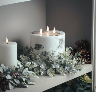 Close-up of the frosted eucalyptus wreath arranged with two LED candles and pinecones on a light grey shelf.