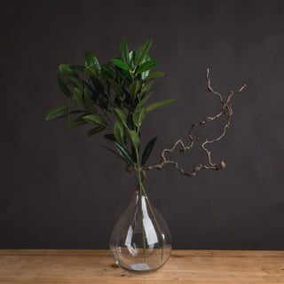Faux bay leaf spray in a clear glass vase on a wooden surface, styled with a twisted natural branch.