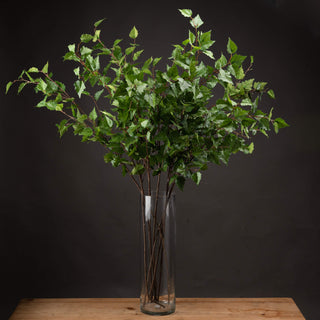 Full view of artificial green branch sprays arranged in a tall clear glass cylinder vase on a wooden surface.