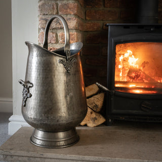 Grey metal bucket on a stone hearth next to a lit wood-burning stove with exposed brickwork.