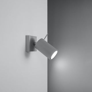 Grey cylindrical wall spotlight fixture mounted in a corner, switched on and casting a bright beam of light onto the adjacent wall.