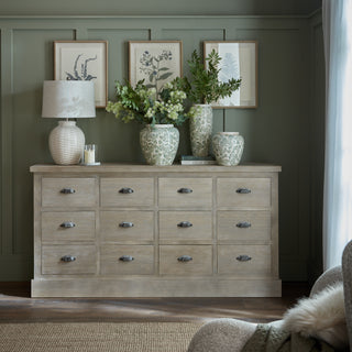 Styled room scene showing the 12 drawer chest in light wood finish against a green panelled wall.
