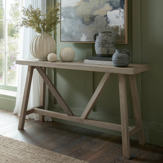 Light wood console table featuring A-frame legs and diagonal support, styled in a room with a green panelled wall.