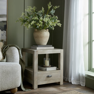 Washed oak side table next to a sofa, showing the single drawer and open storage shelf.