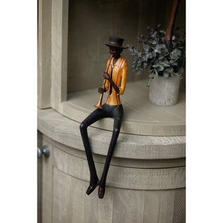 Figurine sitting on a curved wooden shelf next to a grey potted plant, playing a brass instrument.