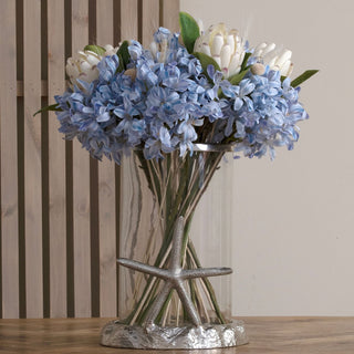 Artificial light blue agapanthus stems mixed with white flowers in a glass vase with a silver starfish decoration.