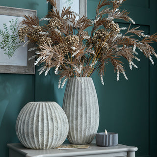 Light grey ceramic vase filled with dried foliage, displayed next to a spherical vase on a table against a teal wall.