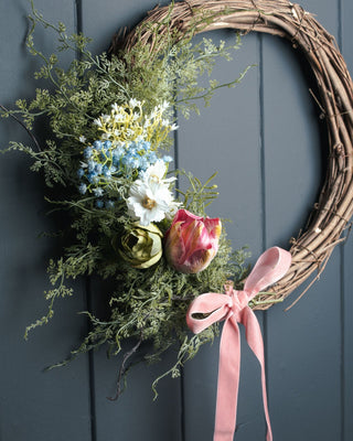 Artificial asparagus fern foliage on a decorative wreath with faux flowers and a pink ribbon, hanging on a dark panelled door.