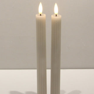 Two taupe ribbed taper candles standing upright against a light background, both illuminated with a warm glow.