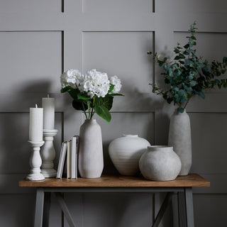 Four coordinating matt white ceramic vases displayed on a wooden table with books and candlesticks against a grey wall.