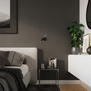 AXIL black wall light fixture installed next to a grey upholstered bed and small metal bedside table.