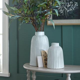 Two ribbed white ceramic vases on a grey console table against a teal panelled wall, filled with faux foliage.