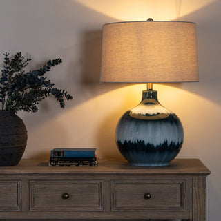 Illuminated table lamp featuring a round blue and white glazed base and a neutral linen shade, displayed on a wooden console.