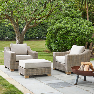 Outdoor footstool with beige cushion placed between two matching rattan armchairs on a garden patio.
