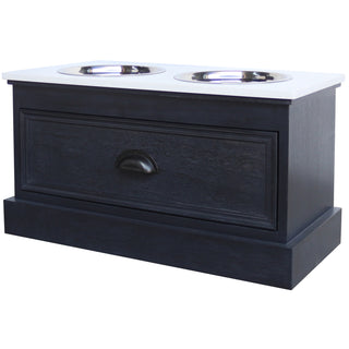 Elevated pet feeder table in dark wood with a white top, featuring two stainless steel bowls and a decorative drawer front.