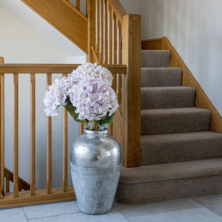 Artificial pink hydrangeas in a large silver floor vase next to an oak staircase with carpeted steps.