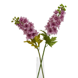 Two artificial purple delphinium stems displayed in a narrow glass vase on a white background.