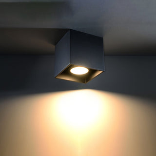 Black cube ceiling light fixture switched on, casting a warm, bright spotlight onto the wall below.