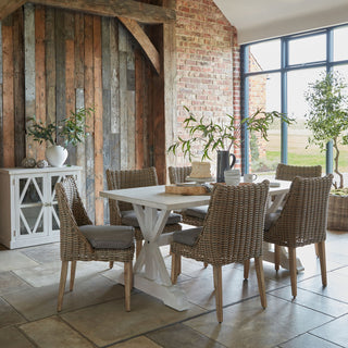 Six woven rattan dining chairs with grey cushions around a white table in a rustic dining room setting.