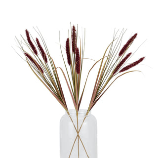 Artificial ruby grass stems displayed fanned out in a clear glass vase against a white background.