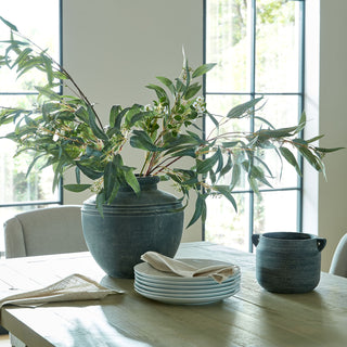 A large arrangement of eucalyptus and mixed foliage in a dark vase on a dining table with plates.