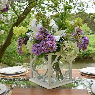 Artificial green hydrangea mixed with purple flowers in a white lantern vase on an outdoor dining table beside a river.