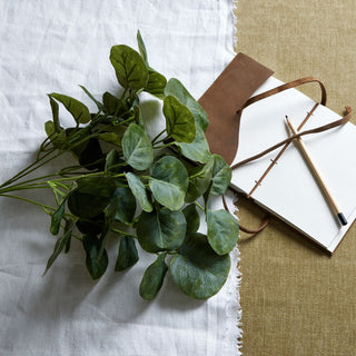 Artificial eucalyptus bunch lying on white fabric next to an open notebook and pencil on a textured khaki surface.